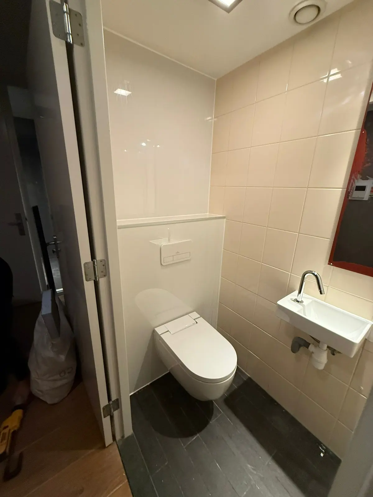 Nieuw gerenoveerd toilet met wit hangtoilet, compact fonteintje en donkere vloertegels. Lichte wandtegels en strak design.