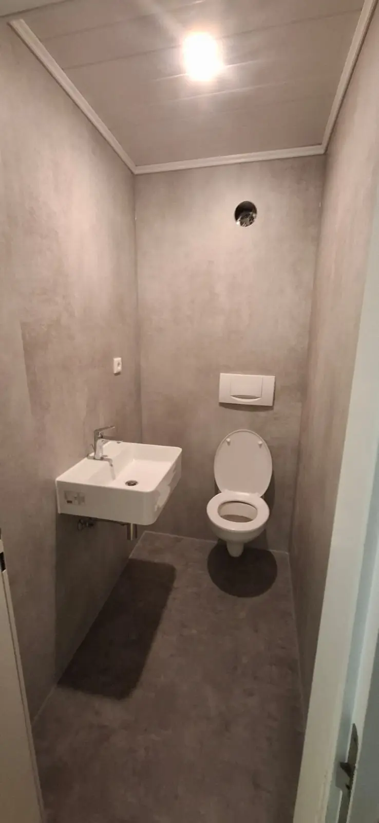 Overzicht van een recent gerenoveerde toiletruimte met grijze wanden, donkere vloer, een wandcloset en een compacte wastafel met kraan.