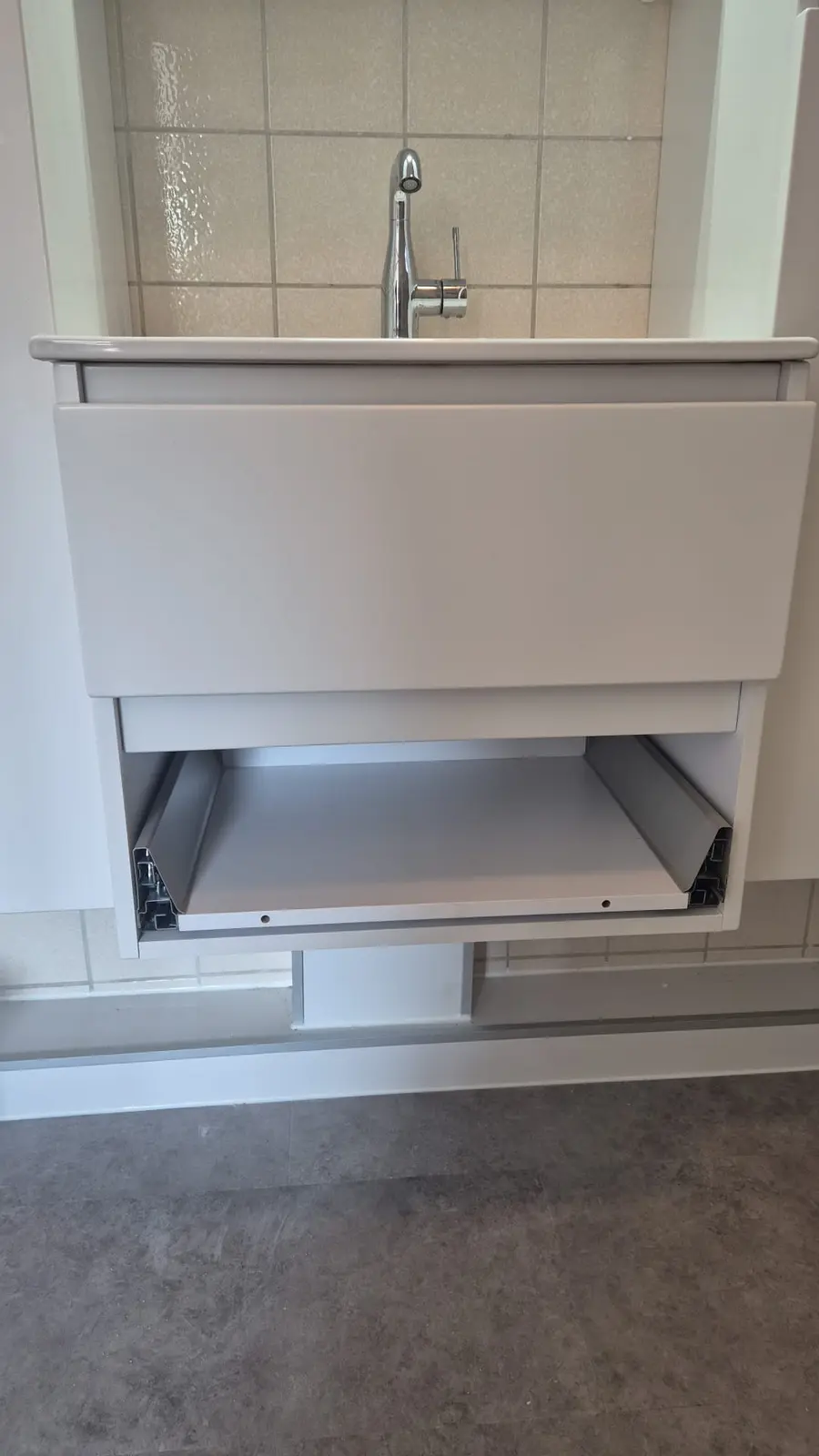 Detail van een moderne witte wastafelonderkast met open lade en chromen kraan in een badkamer met grijze vloer en lichte wandtegels.